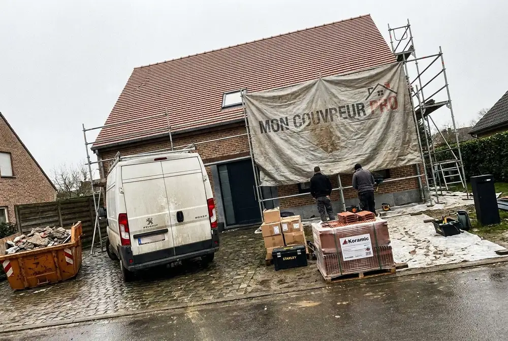 Maison en brique en chantier : échafaudage, bâche Mon Couvreur Pro, camionnette, conteneur et palettes de tuiles Koramic